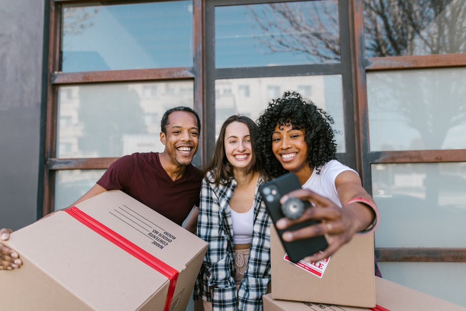 Three individuals standing outdoors in front of a building with large windows, smiling as they pose for a selfie. The person on the right, a woman with curly black hair wearing a white t-shirt, holds