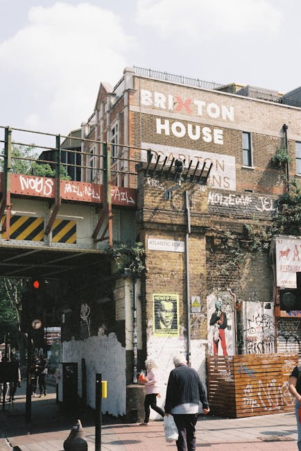 A city street scene showing a brick building with multiple layers of exterior walls, featuring large painted signs including the words 'BRIXTON HOUSE' and various graffiti and posters. A metal fire es