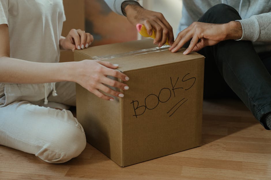 Close-up of two individuals packing a cardboard box labeled 'BOOKS' in black marker, with one person holding the box steady while the other uses a yellow tape dispenser to seal it. The box is placed o