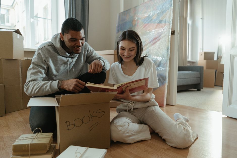 Close-up of two individuals packing a cardboard box labeled 'BOOKS' in black marker, with one person holding the box steady while the other uses a yellow tape dispenser to seal it. The box is placed o