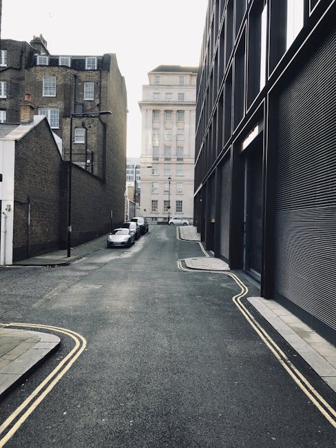 A narrow urban street scene showing a steep, one-way road with double yellow lines along the curb edges. On the left side, there are old brick buildings with multiple windows and rooftop structures, w