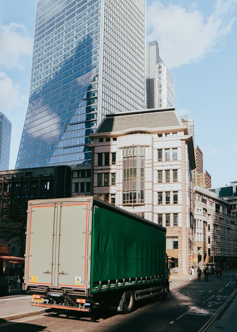 A large green moving truck with closed rear doors is parked on a city street in front of tall modern office buildings with glass facades and older architectural structures. The truck is positioned nea